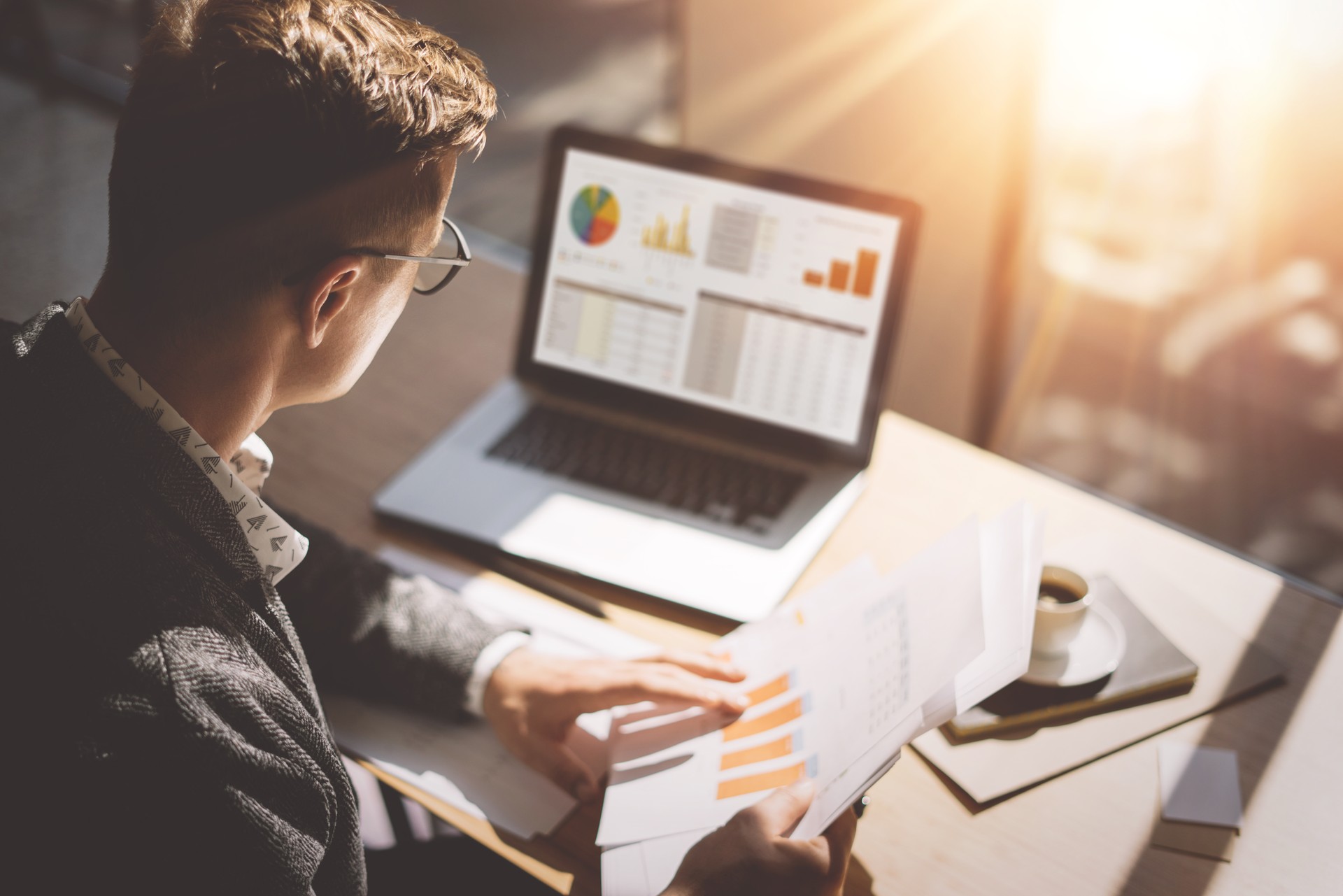 Jeunes des Finances analyste de marché à lunettes travaillant au bureau ensoleillé sur ordinateur portable tout en étant assis à une table en bois. Homme d’affaires analyser le document dans ses mains. Graphes et schéma sur l’écran de l’ordinateur portable. Floue.